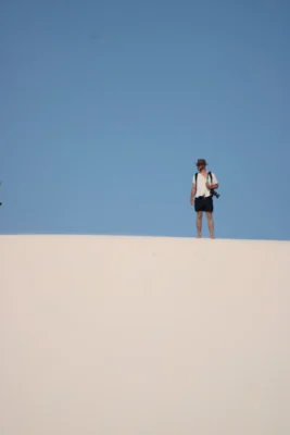 Viajante solo de mochila e chapéu parado no alto de uma duna branca dos Lençóis Maranhenses